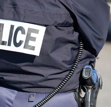 The back of the police officer with the inscription "POLICE" with a gun and a walkie-talkie. Découverte macabre au Grau-du-Roi : un adolescent avoue le meurtre