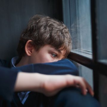Pensive sad boy teenager in a blue shirt and jeans sitting at the window and closes his face with his hands. Soutien pour les Enfants Souffrant de Phobie Scolaire