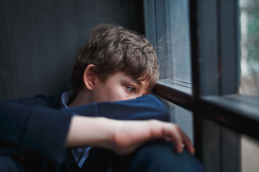 Pensive sad boy teenager in a blue shirt and jeans sitting at the window and closes his face with his hands. Soutien pour les Enfants Souffrant de Phobie Scolaire